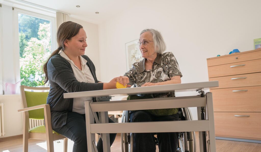 Ergotherapeutin unterstützt ältere Patientin bei feinmotorischer Übung am Tisch.
