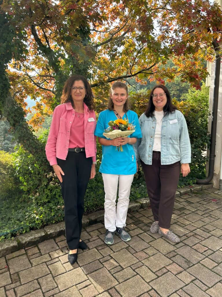 Lucie Tschudy (Mitte) mit einem Blumenstrauß, begleitet von zwei Kolleginnen der Winkelwaldklinik, anlässlich ihrer erfolgreichen Anerkennung als Pflegefachfrau in Deutschland.