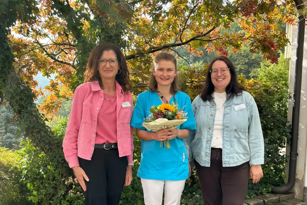 Lucie Tschudy (Mitte) mit einem Blumenstrauß, begleitet von zwei Kolleginnen der Winkelwaldklinik, anlässlich ihrer erfolgreichen Anerkennung als Pflegefachfrau in Deutschland.