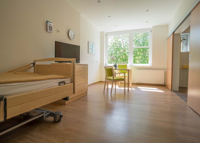 Großzügiges Patientenzimmer mit Pflegebett, Fernseher, Sitzbereich und großem Fenster mit Blick auf Bäume.